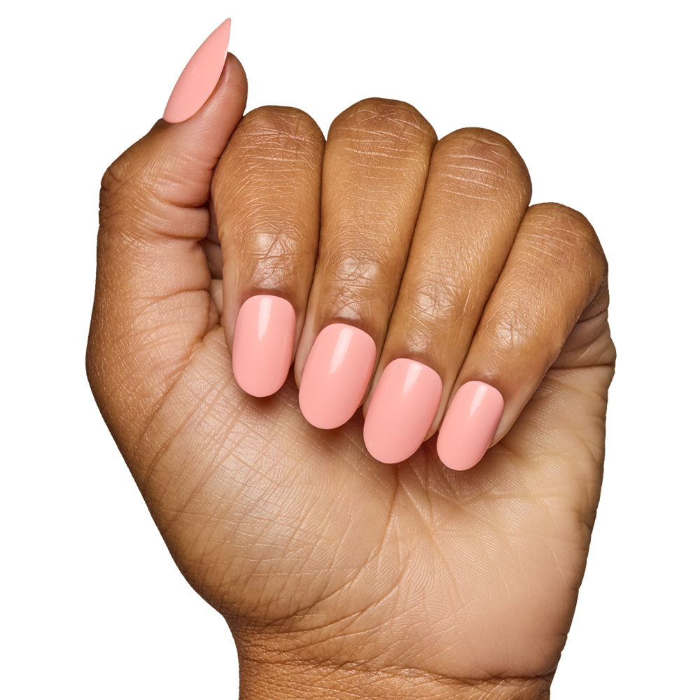 A hand with medium brown skin displays almond-shaped nails coated in glamnetic’s Peach Coral, a glossy pastel peach shade. The pinky nail is longer and more pointed, echoing the elegance of the Peach Coral Nail Set.