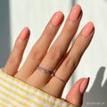 A hand with neatly manicured, oval nails painted in glamnetics Peach Coral shade. Two silver rings decorate the middle finger, and a yellow-and-white striped sleeve partially covers the wrist.