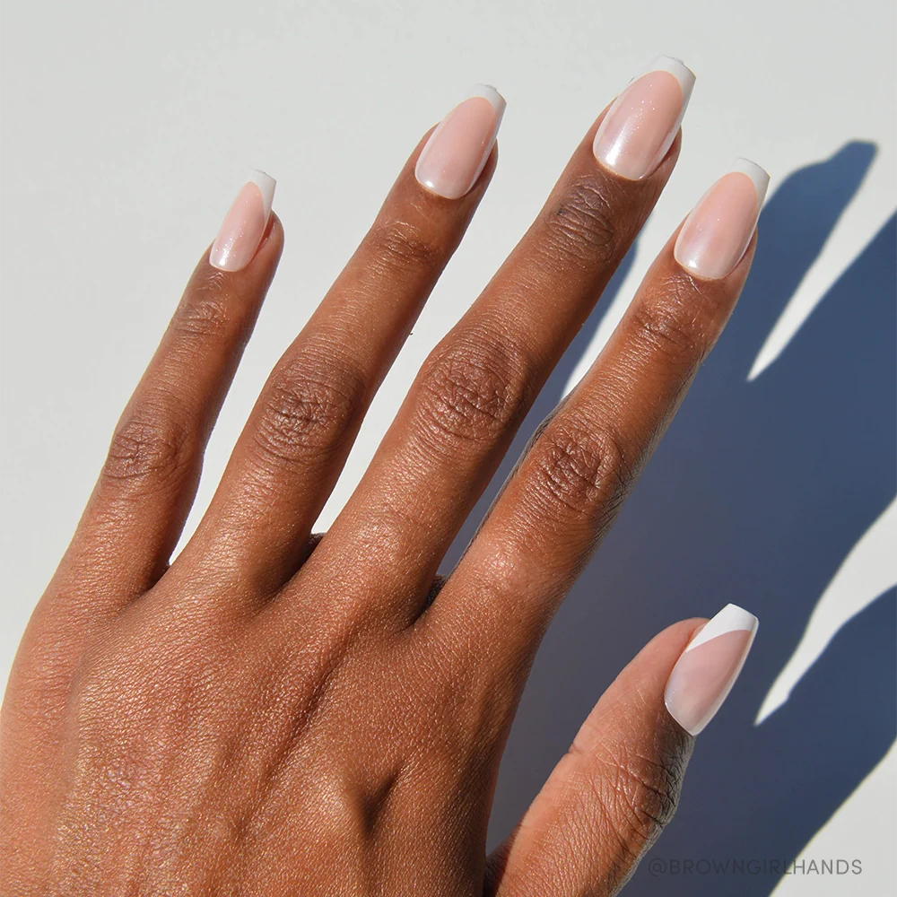 A hand with medium brown skin displays neatly manicured nails in glamnetic's Pearl Glaze, featuring a subtle French tip and medium coffin shape, set against a white background with visible shadows.