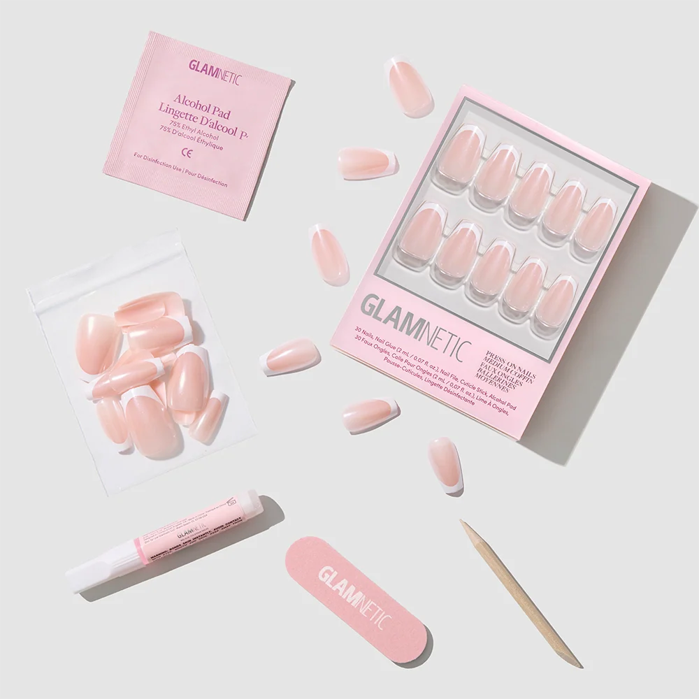 A flat lay shows the glamnetic Pearl Glaze Press-On Nails kit with medium coffin pink nails, extra nails, an alcohol pad, glue, pink file, and a wooden cuticle stick on a light grey background.