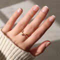 A hand with a light skin tone and a French tip manicure wears the glamnetic "Pearly Please" gold ring adorned with small gemstones. The beige ribbed sweater and softly blurred background enhance the look’s elegance.