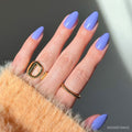A hand with long, almond-shaped glamnetic Periwinkle press-on nails, glossy and periwinkle blue, wearing gold rings and partly covered by a fuzzy beige sweater sleeve. The softly lit background is plain.