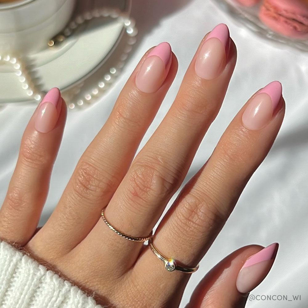 A hand with an almond base wears glamnetics Pink Macaron nails in pink French tips. Two delicate gold rings adorn the fingers, and a cup with a pearl necklace appears in the background.