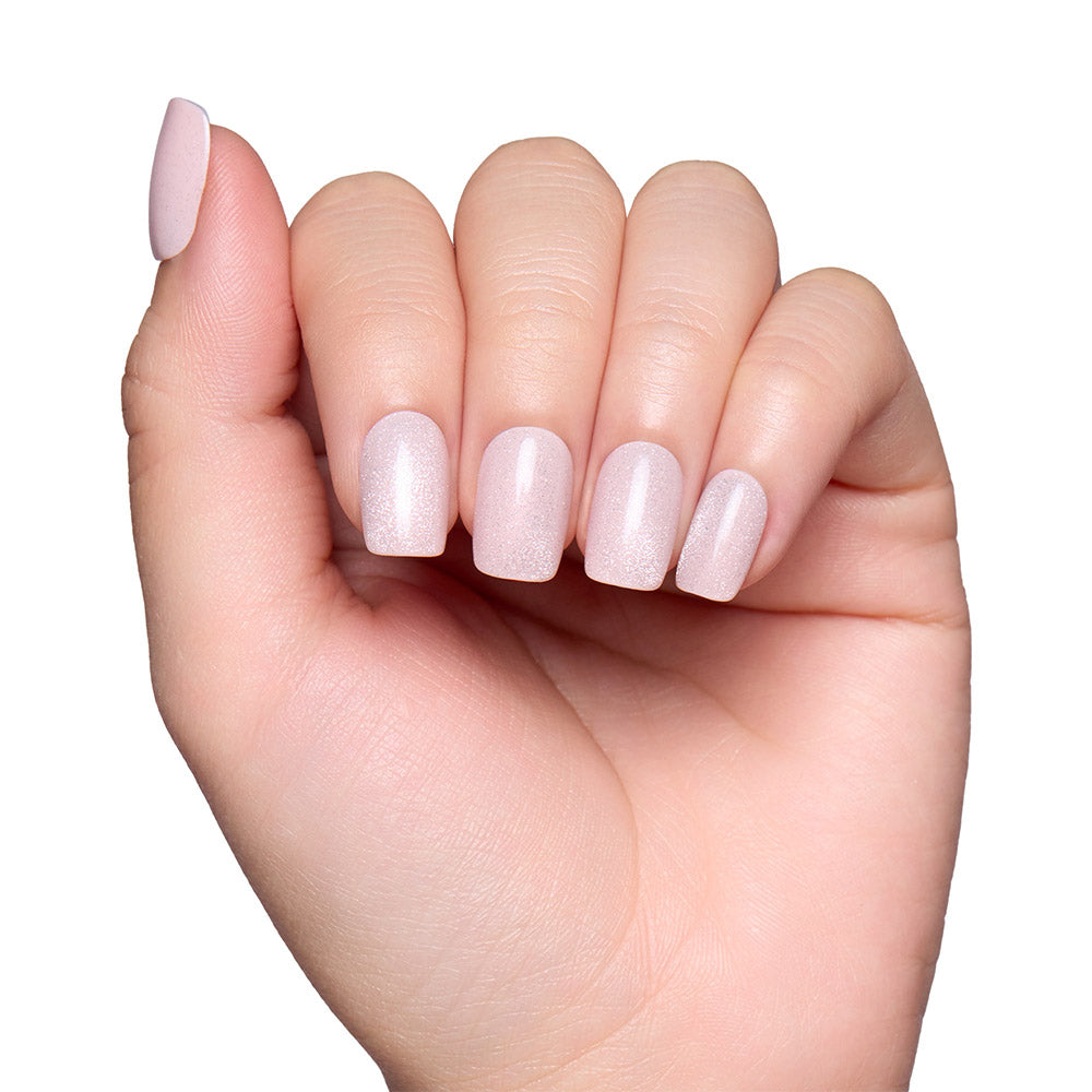 A hand with short squoval nails is painted in glamnetics Pinky Promise—a pale, shimmery pink polish with a blush velvet finish—shown against a white background.