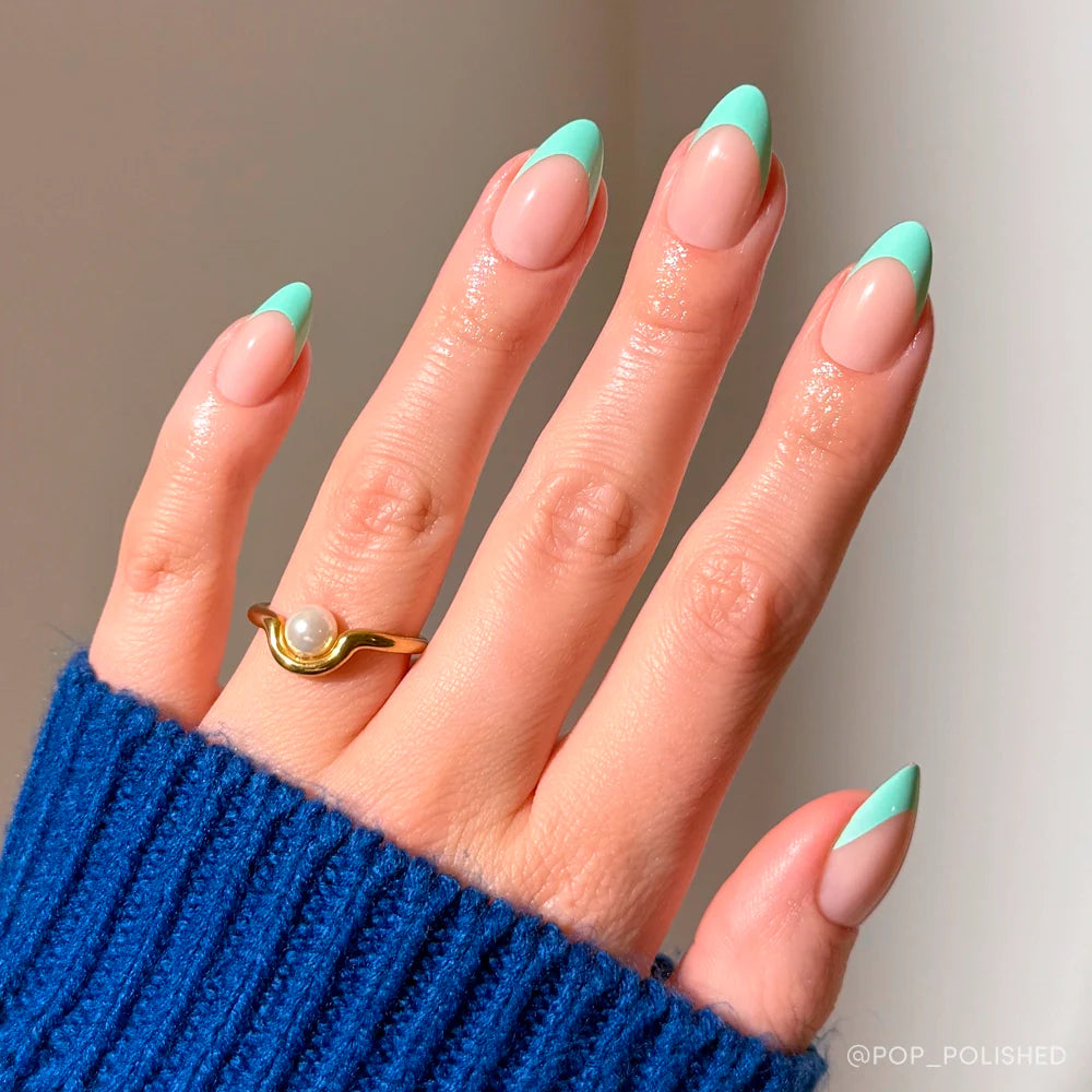 A hand with glamnetic Pistachio Crème almond-shaped French tip press-on nails in mint green is shown. The person wears a blue sweater and a gold pearl ring, with soft lighting highlighting the salon-quality nails against a neutral backdrop.