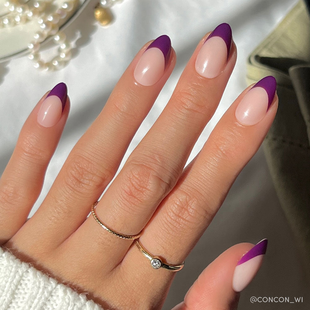 A hand with short almond nails in glamnetic’s Plum features a nude base and dark purple French tips for a chic look. Two gold rings adorn the fingers as sunlight highlights the nails, with pearls and fabric softly blurred in the background.