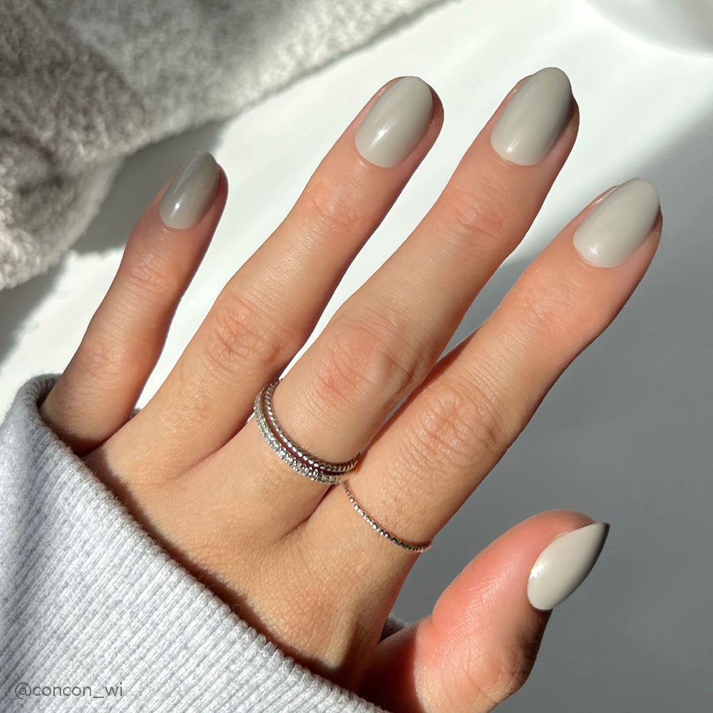 A hand with neatly manicured Pure Gray nails by glamnetic, featuring a glossy finish, rests on a white surface. The person wears a light gray sweater and two silver rings on the middle finger as sunlight casts soft shadows.