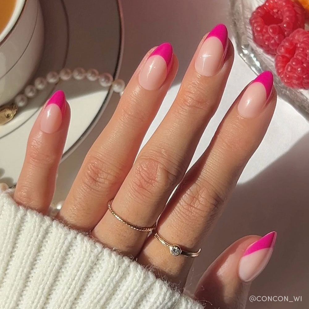 A hand with almond nails sports glamnetics Raspberry Sorbet glossy magenta-pink French tips, paired with two delicate gold rings and a white sweater. Pearls, tea, and raspberries are seen in the background.
