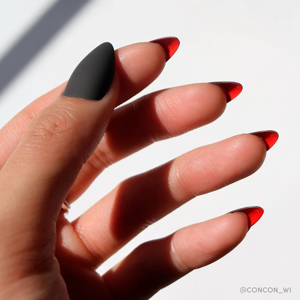 A hand displays glamnetics Red Bottom press-on nails—long stilettos with a red, translucent ombre on four fingers and a matte black thumb—highlighting their luxurious style against a white background.