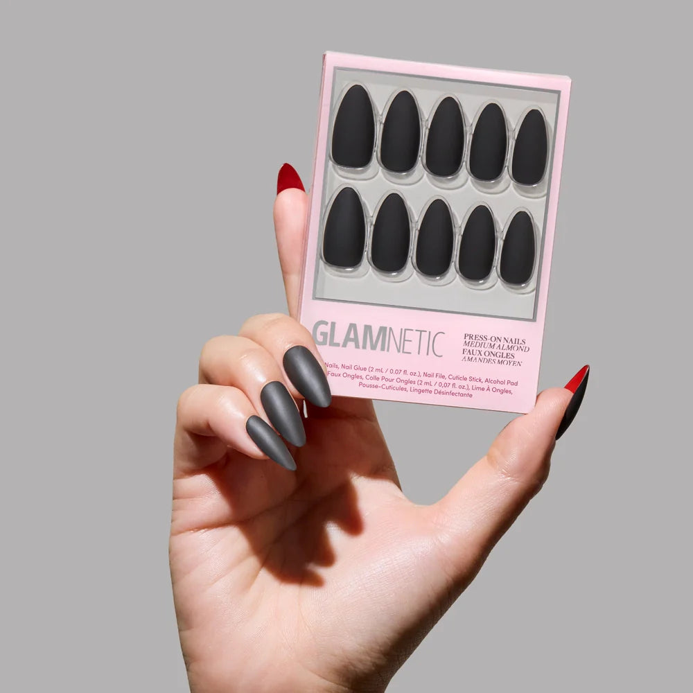 A hand with long, black matte almond nails from glamnetic’s Red Bottom set holds the box against a plain grey background. The thumb nail reveals a bold red underside.