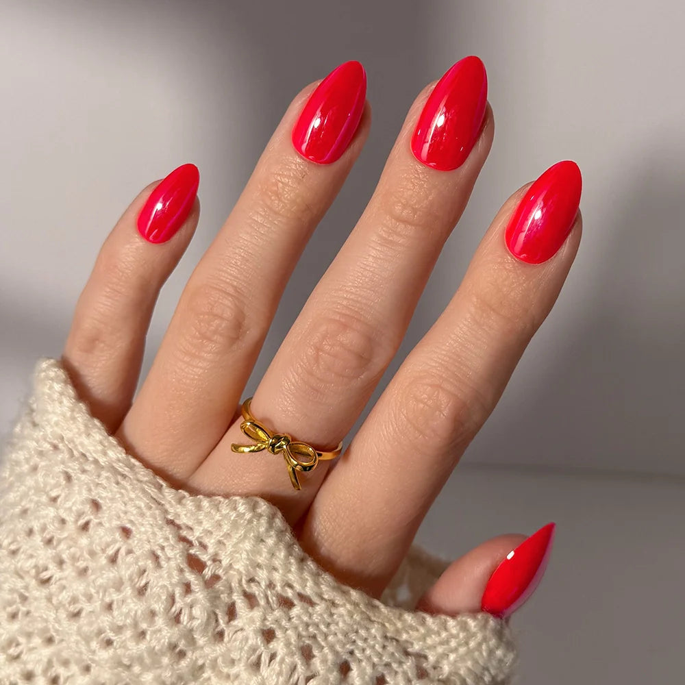 A hand with long, glazed red nails wears a gold bow ring. Styled in a cream knit sleeve, the elegant vegan, cruelty-free "Red Flag" press-on nails by glamnetic are softly lit in the background.
