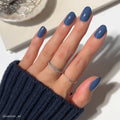 A hand with neatly manicured nails painted in Regal Blue matte polish, wearing two delicate silver rings and a navy ribbed sweater. The scene includes a white surface and some jewelry in the background.