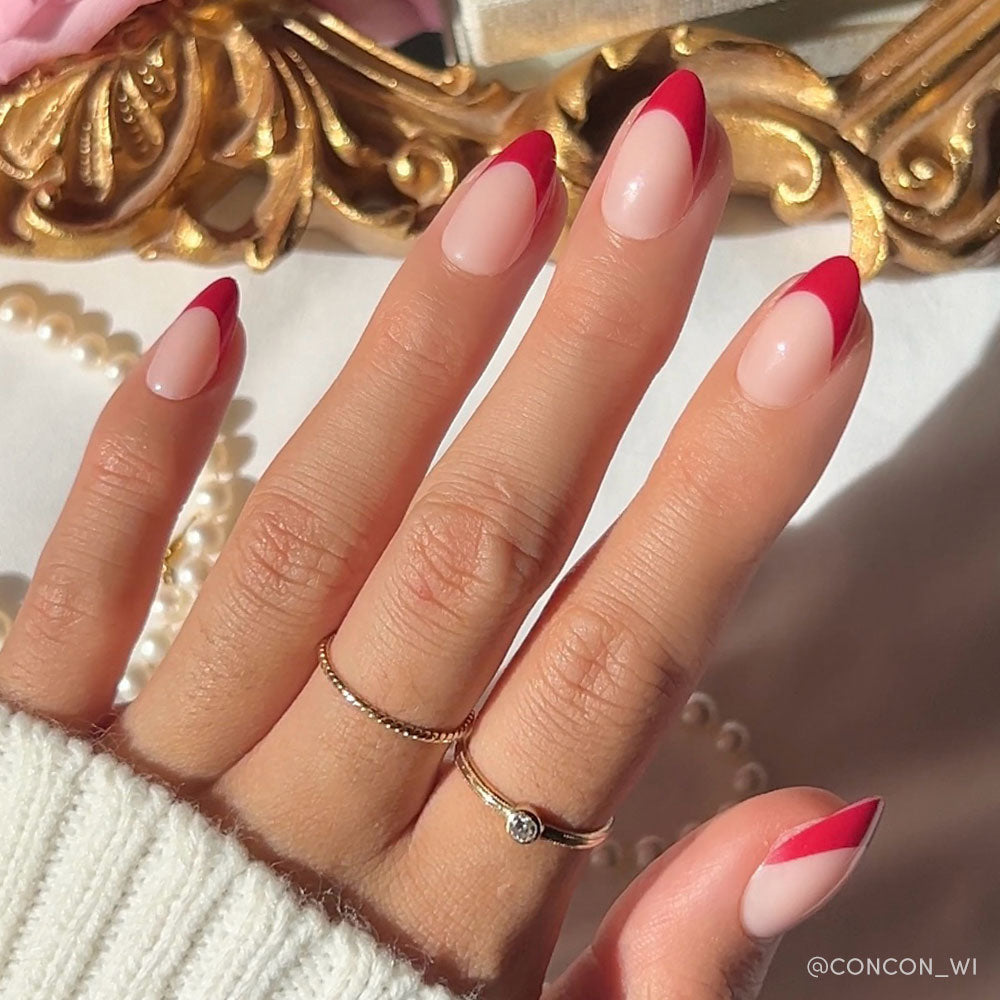 A hand with short almond nails from glamnetics Rose Garden set shows a pink base and deep red French tips. Two gold rings shine, paired with a white sweater sleeve and pearl necklace for an elegant finish.
