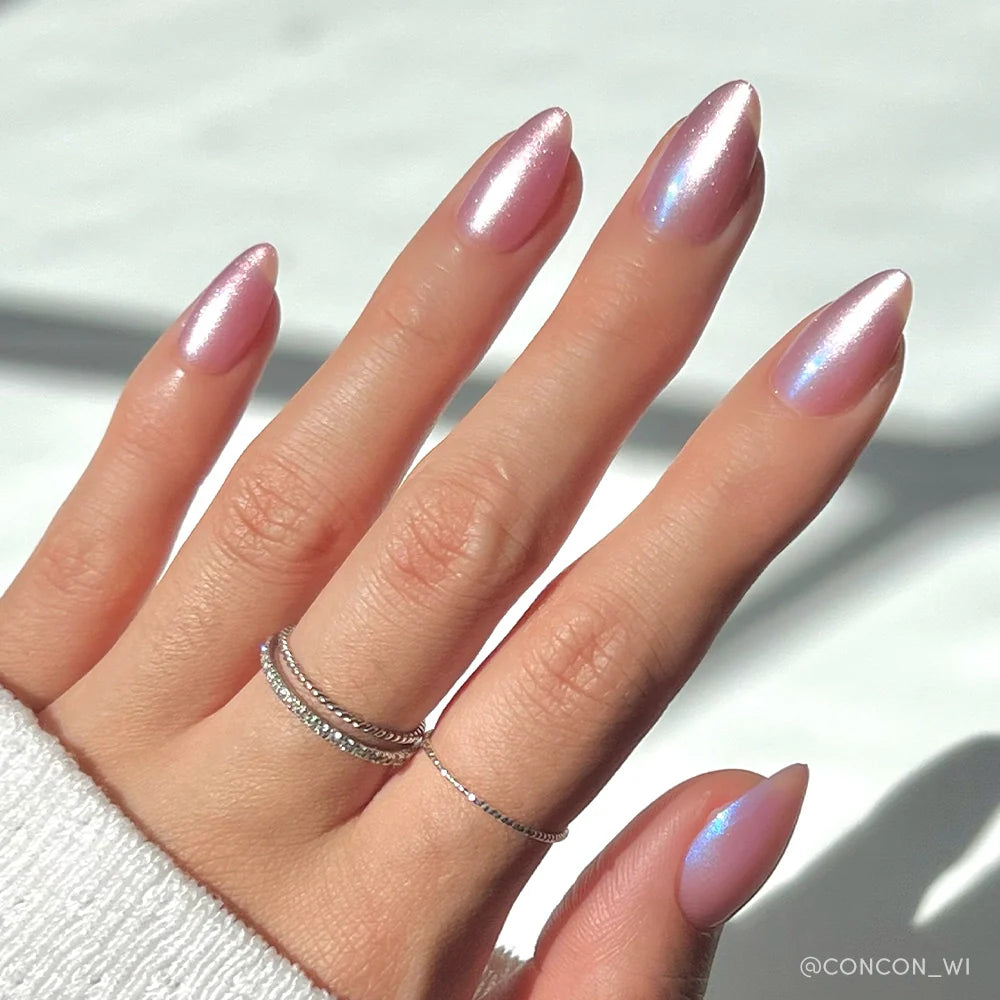 A hand with short almond nails from glamnetic’s Seaspell set, painted in a shimmering velvet light pink. Three delicate silver rings adorn the fingers, and a white sweater sleeve partially covers the wrist.
