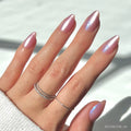 A hand with short almond nails from glamnetic’s Seaspell set, painted in a shimmering velvet light pink. Three delicate silver rings adorn the fingers, and a white sweater sleeve partially covers the wrist.