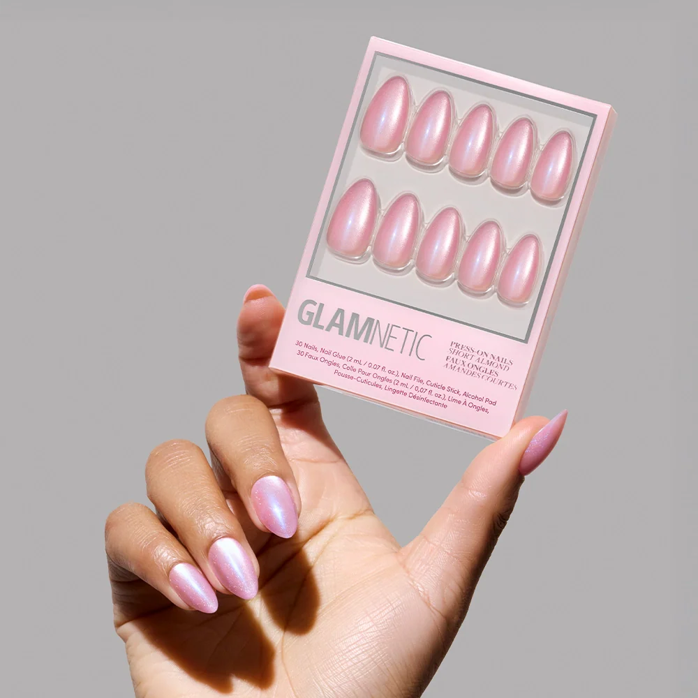 A hand with iridescent pink short almond nails holds a glamnetic Seaspell Press-On Nail Set in a pearly pink, shimmering velvet finish against a neutral gray background.