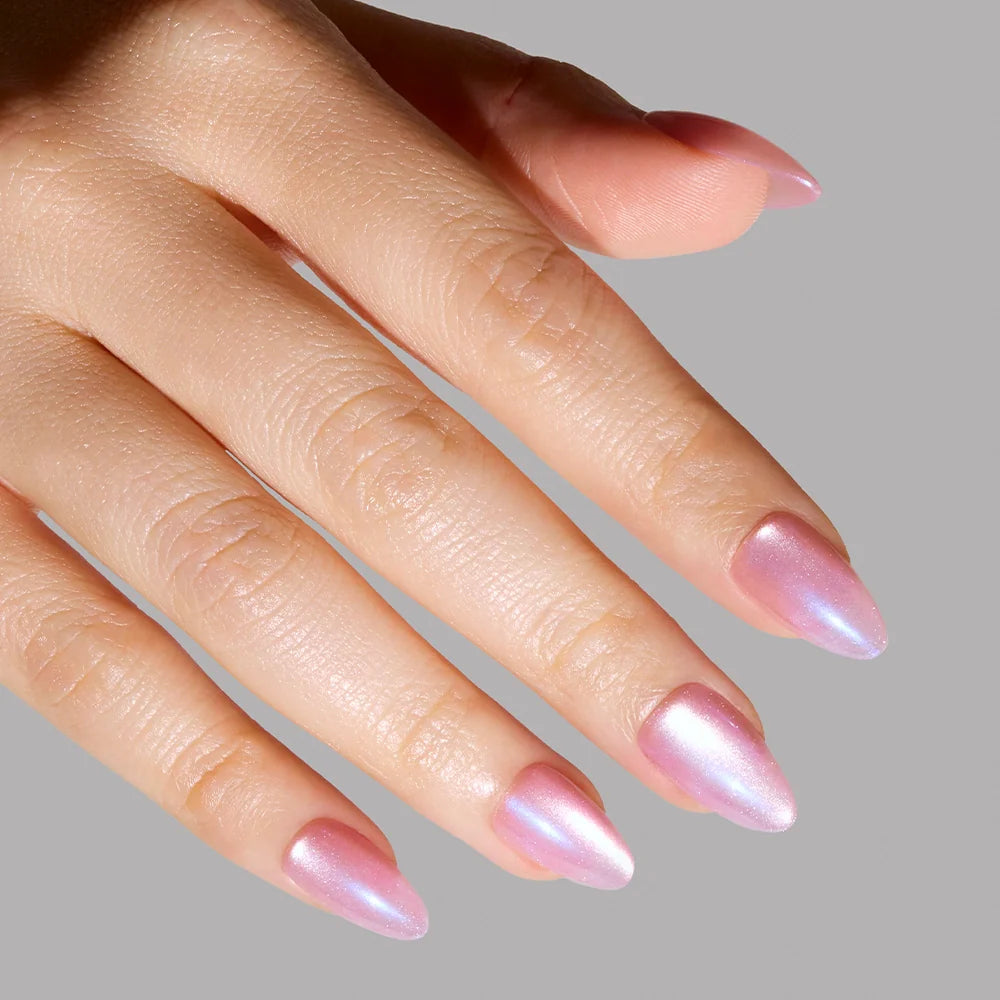 A hand with short almond nails is painted in glamnetics Seaspell, an iridescent pink polish with a shimmering velvet finish, shown against a plain gray background.
