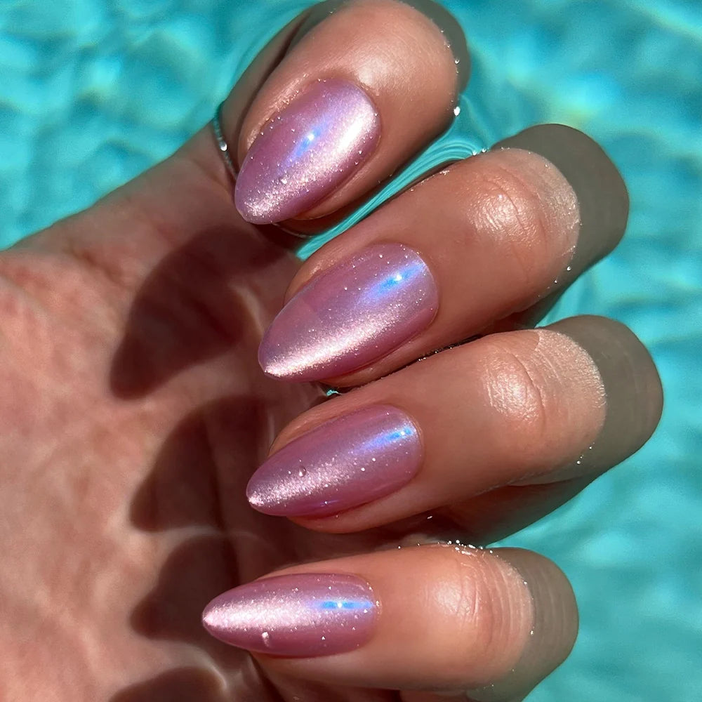 A hand with almond-shaped nails in an iridescent pink velvet finish showcases the glamnetic Seaspell Press-On Nail Set by a clear blue pool, evoking a bright, sunlit outdoor vibe.