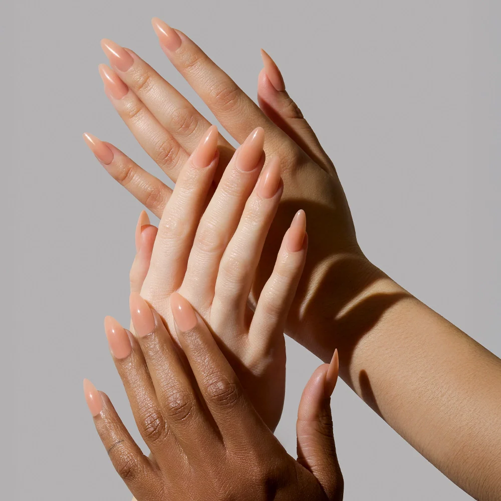 Three hands with different skin tones overlap, showcasing long, almond-shaped nails painted in glamnetic’s Sheer Peach vegan & cruelty-free polish—a semi-transparent peachy nude jelly—against a light grey background.