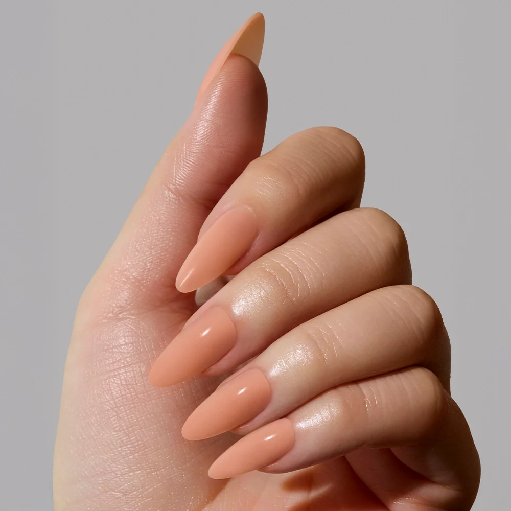 A hand with long, almond-shaped nails painted in glamnetics Sheer Peach, a smooth matte nude shade, is shown against a plain light gray background.