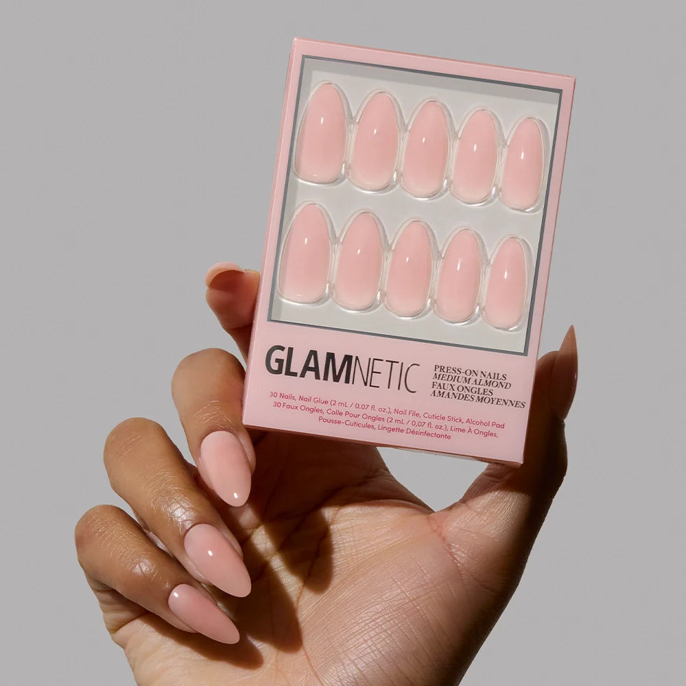 A hand holds a box of glamnetic Sheer Pink press-on nails, while the other shows several applied in the same shade. The vegan, cruelty-free set includes ten glossy, almond-shaped nails on neutral packaging.