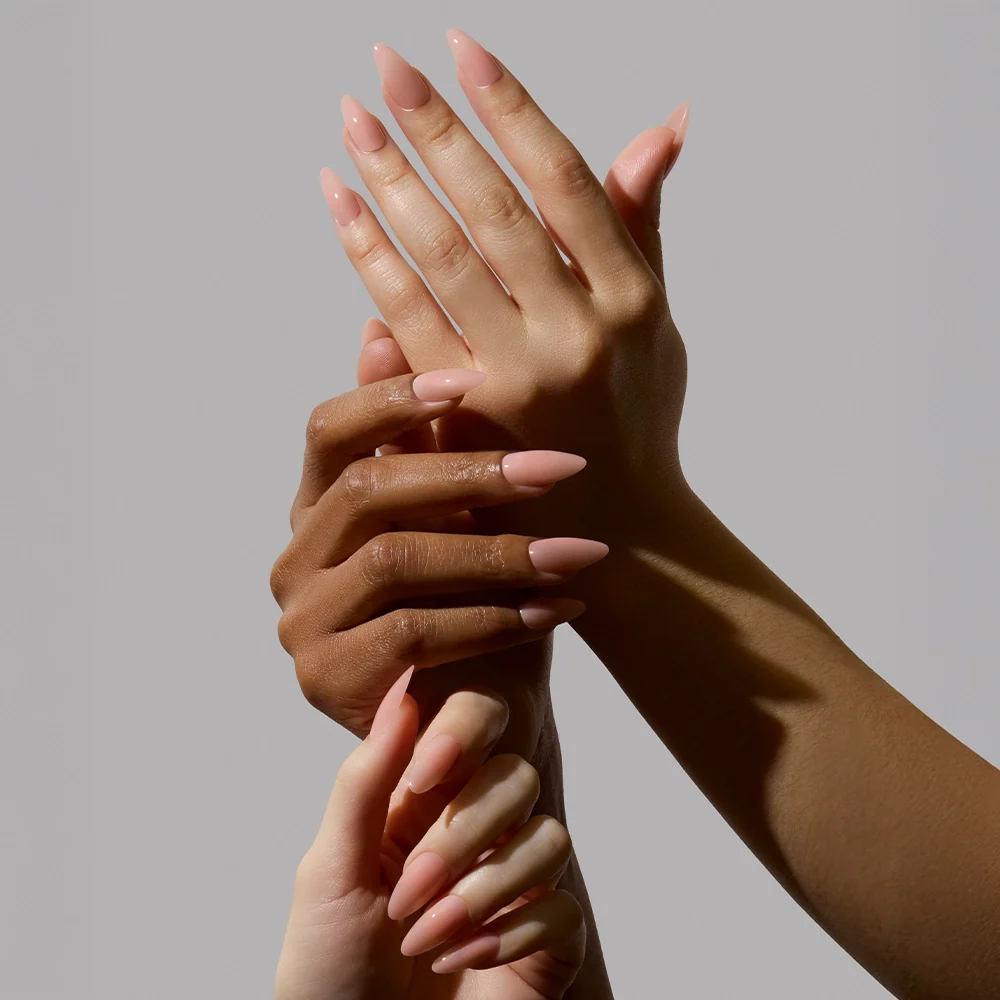 Three hands with different skin tones display glamnetics Sheer Pink nails—long, pointed, and nude-pink—intertwined against a plain gray background. This image highlights diversity and the perfect soft-glam look for a clean-girl aesthetic.