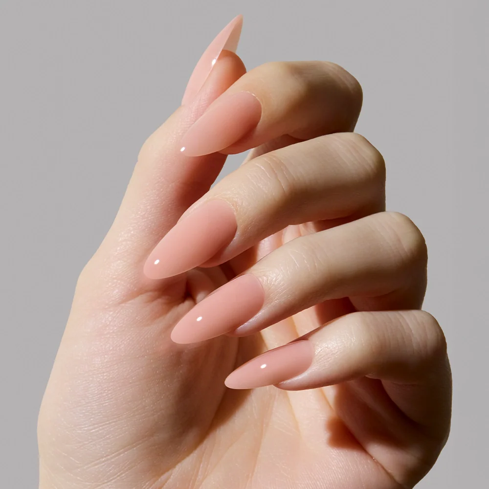 A hand with long, almond-shaped nails painted in glamnetics Sheer Pink polish—a glossy nude pink shade perfect for a clean-girl look—displayed against a plain, light gray background.