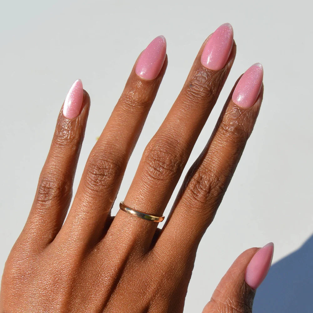 A hand with medium brown skin wears a simple gold ring and displays long nails painted in glossy rosy pink velvet—perfectly showcasing glamnetic’s Shine for You nail set against a light background.