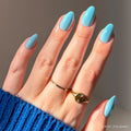 A hand with short almond-shaped nails painted in glamnetics Soft Blue, wearing two gold rings—one plain, one round—paired with a blue knit sweater sleeve.