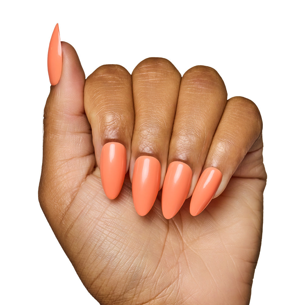 A hand with medium brown skin displays long, almond-shaped nails in glamnetic’s Soft Tangerine—a salon-quality acrylic look with a glossy pastel orange finish—set against a white background.