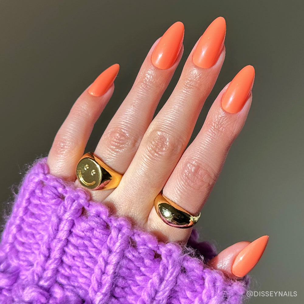 A hand with long, almond-shaped nails painted in Soft Tangerine by glamnetic, adorned with two gold rings—one featuring a smiley face—rests on a chunky, purple knitted sweater.
