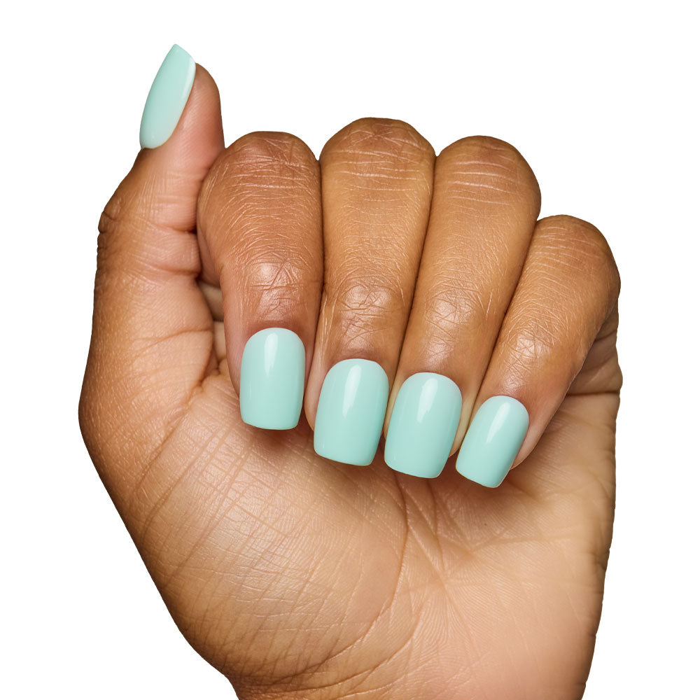 A hand with medium brown skin shows short squoval nails painted in glamnetic’s Soft Teal set. The loose fist pose against a white background highlights the glossy, fresh manicure.