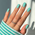 A hand with neatly manicured short squoval nails painted in glamnetics Soft Teal. Thin silver rings adorn the fingers, and a green-and-white striped sleeve partially covers the wrist as bright sunlight casts soft shadows.