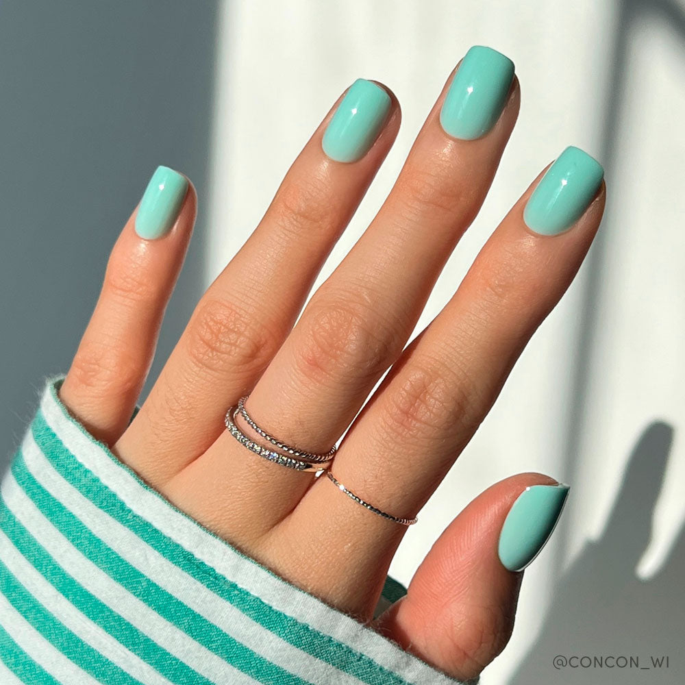 A hand with neatly manicured short squoval nails painted in glamnetics Soft Teal. Thin silver rings adorn the fingers, and a green-and-white striped sleeve partially covers the wrist as bright sunlight casts soft shadows.