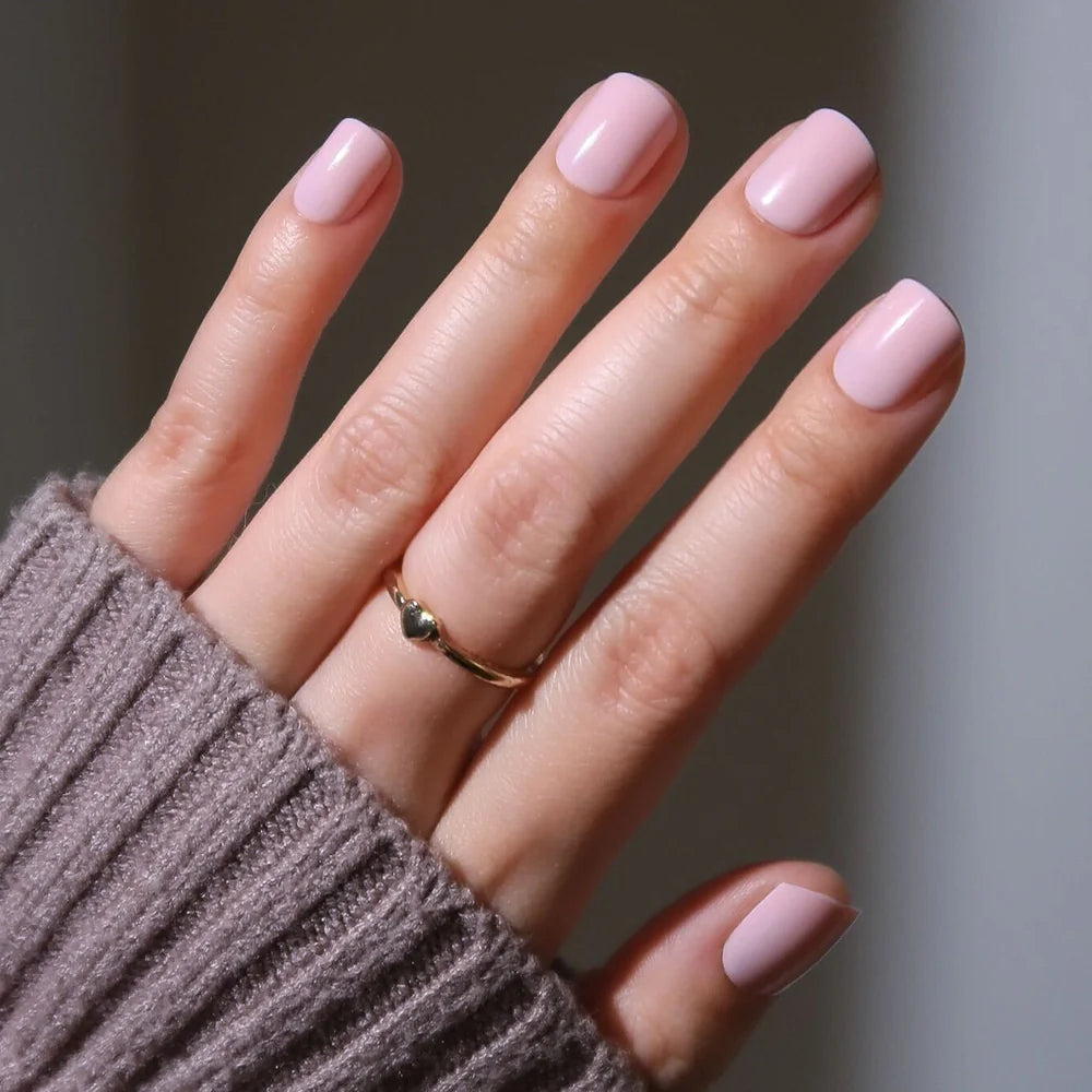 A hand with super short nails painted in glamnetic’s Sugar BB classic pink, wearing a simple gold heart ring and a mauve ribbed sweater sleeve partially covering the wrist.