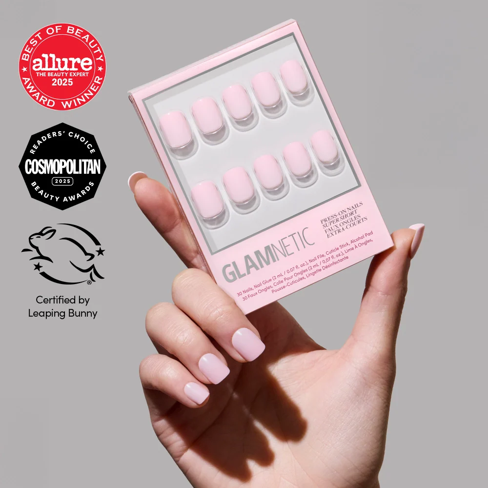 A hand with a classic pink mani of glossy Sugar BB press-on nails by glamnetic holds a box of similar super short nail press-ons. The image displays Allure, Cosmopolitan, and Leaping Bunny certification logos.