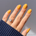 A hand with glossy, citrusy Sunset Orange nails by glamnetic wears a large emerald-cut diamond ring. Paired with a dark blue ribbed sweater and bright natural light, this look brings a fresh touch to summer style.