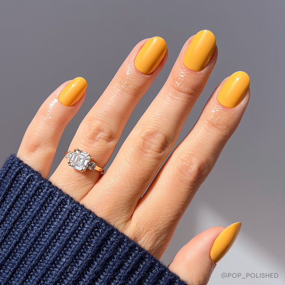A hand with glossy, citrusy Sunset Orange nails by glamnetic wears a large emerald-cut diamond ring. Paired with a dark blue ribbed sweater and bright natural light, this look brings a fresh touch to summer style.