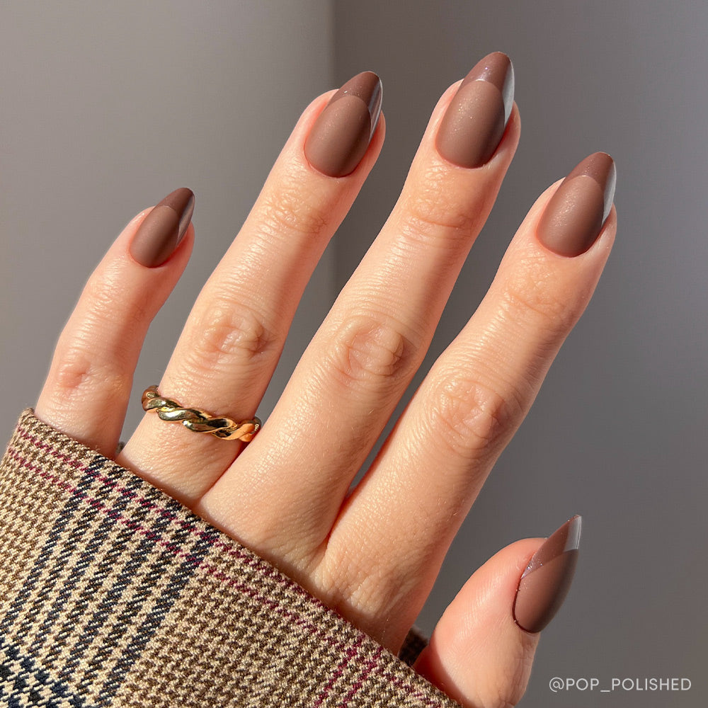 A hand with glamnetic Terra almond-shaped nails in earthy brown, featuring a matte finish and glossy brown half-moon French tips, is accessorized with a gold twisted ring and paired with a plaid sleeve.