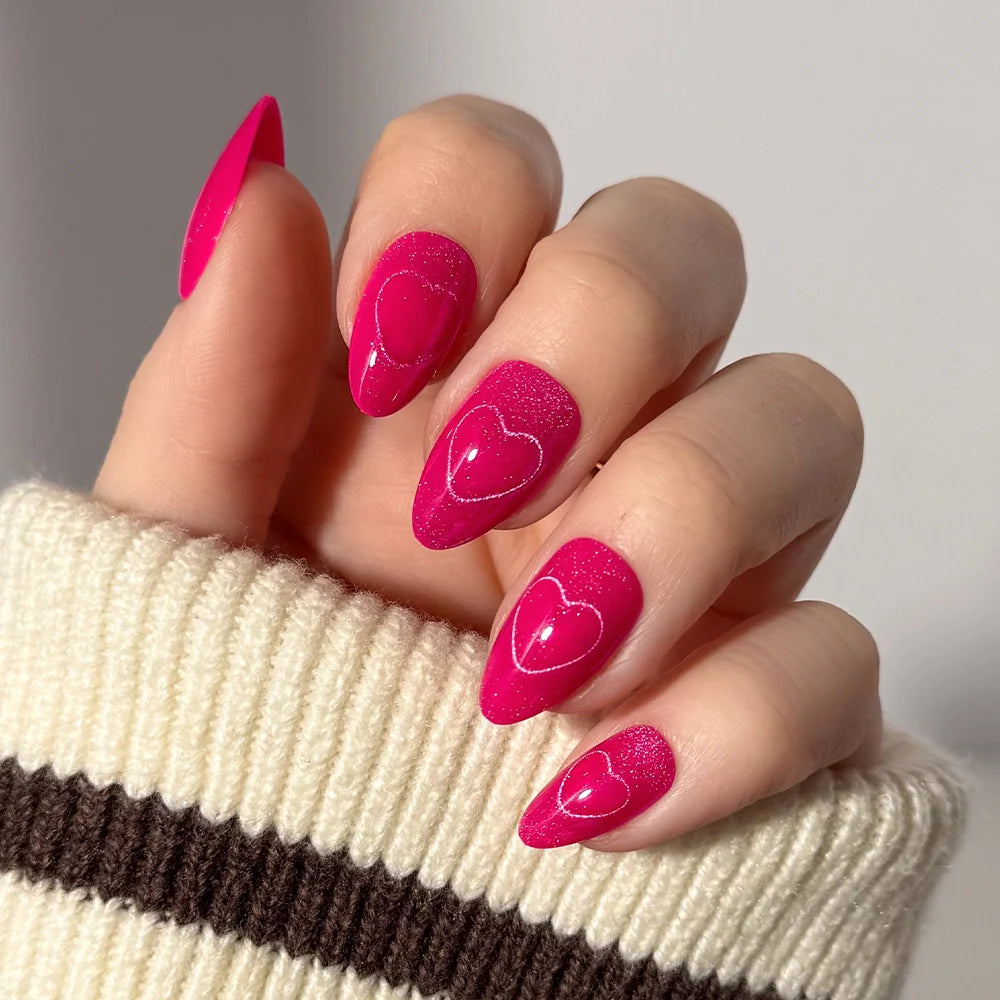 A hand with almond-shaped, bright pink nails featuring glossy heart designs from the glamnetic Text Me Back set. The person is wearing a cream sweater with a brown stripe.