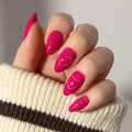 A hand with almond-shaped, bright pink nails featuring glossy heart designs from the glamnetic Text Me Back set. The person is wearing a cream sweater with a brown stripe.