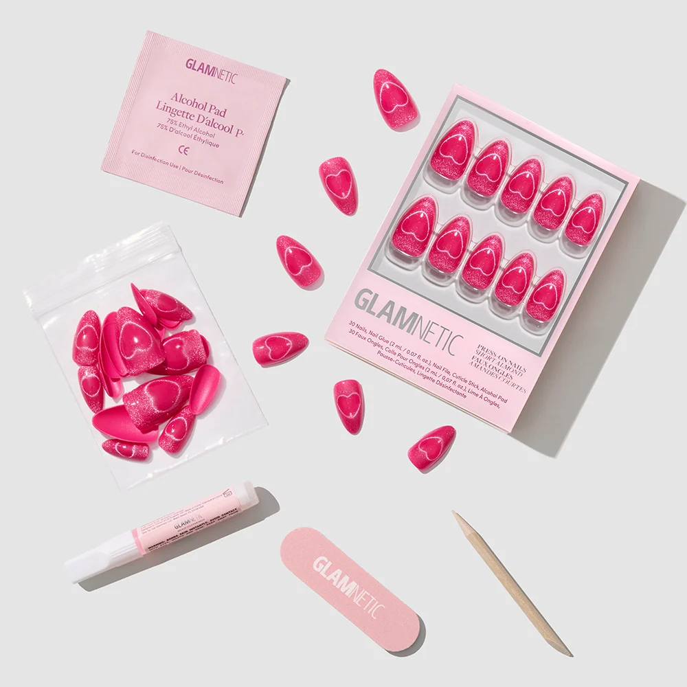 A flat lay of the glamnetic Text Me Back set displays hot pink velvet nails with heart designs, plus an alcohol pad, nail glue, mini file, cuticle stick, and extra nails all neatly arranged on a white background.
