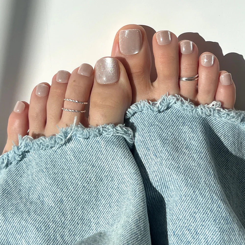 Close-up of feet with shimmering champagne silver Glamnetic press-on toenails, styled with silver rings and light-washed frayed denim jeans.