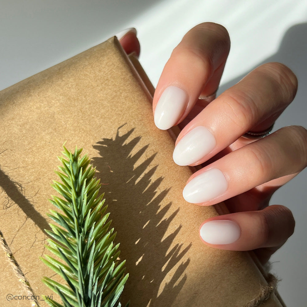 A hand with neatly manicured, short oval nails in glamnetics White Swan shade holds a brown paper-wrapped gift decorated with a pine branch; sunlight highlights the nails’ glossy finish.