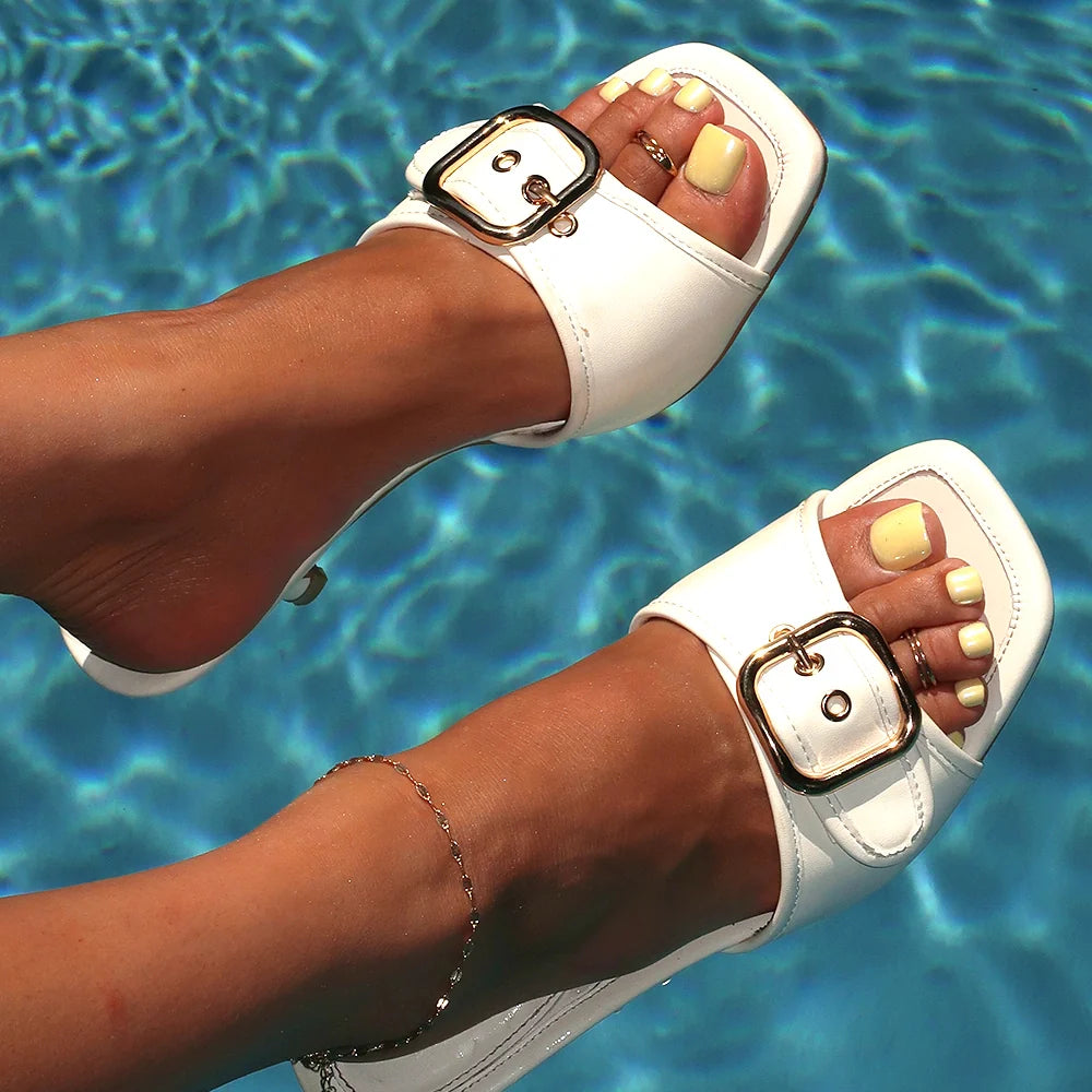Feet with pastel yellow press-on toenails wearing white sandals, posed above a rippling blue pool, accessorized with an anklet and toe rings.