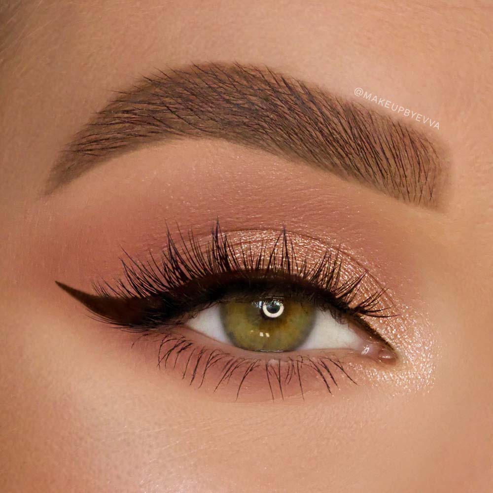A close-up of a green eye with bold winged black eyeliner, shimmery peach eyeshadow, a well-groomed thick eyebrow, and long vegan Babygirl lash extensions by glamnetic.