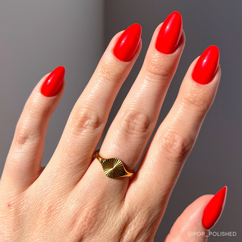 A hand with glamnetic’s Fiery Red almond-shaped nails and a gold sunburst ring on the middle finger pops against a neutral background—perfect for standout summer nails.
